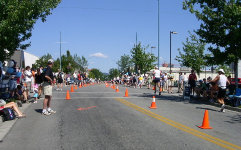 [A two-lane road with double yellow lines in the center has orange cones in the center and along both sides. Plenty of spectators behind the cones are encouraging the few runners currently at this point of the race.]